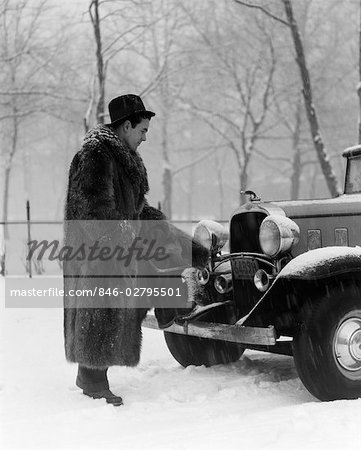 1930s MAN IN HAT AND RACCOON FUR COAT STANDING FOOT ON BUMPER OF CHEVROLET ROADSTER STALLED IN SNOW STORM