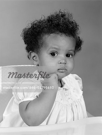 1960s AFRICAN AMERICAN BABY GIRL IN DRESS SITTING IN HIGH CHAIR WITH HAND ON CHIN & SERIOUS EXPRESSION