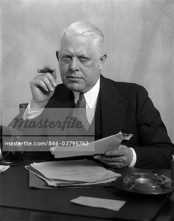 1930s MAN IN OFFICE SITTING AT DESK HOLDING PAPERS SMOKING A CIGAR