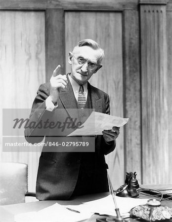 1930s ELDERLY MAN IN OFFICE STANDING BEHIND DESK GESTURING WITH RAISED FINGER