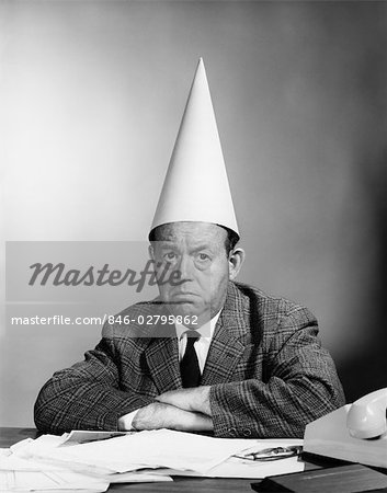 1960s UNHAPPY BUSINESSMAN BEHIND DESK WEARING DUNCE CAP LOOKING AT CAMERA