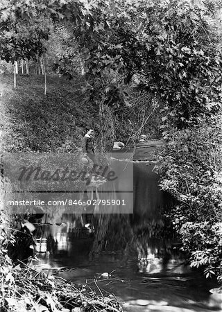 1890s TURN OF THE CENTURY BOY STANDING IN WOODED AREA LOOKING DOWN AT REFLECTION IN CREEK