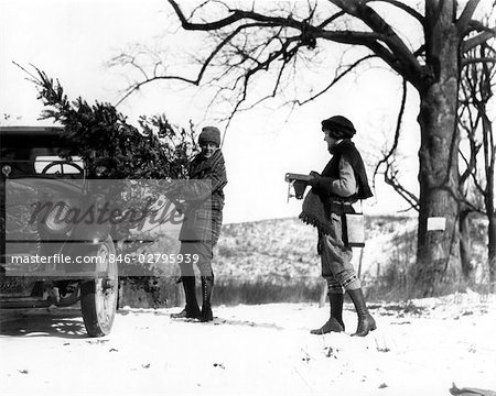 1920s MAN LOADING CHRISTMAS TREE INTO CAR WITH WOMAN HOLDING CAMERA WATCHING HIM