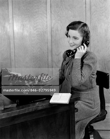 ANNÉES 1940 SOURIANTE JEUNE FEMME SECRÉTAIRE RÉCEPTIONNISTE ASSIS AU BUREAU BUREAU PARLANT ON TÉLÉPHONE