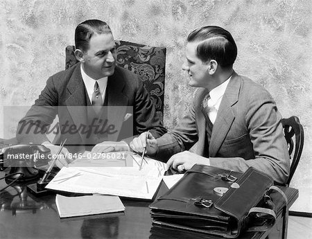1930s TWO MEN SITTING ON SAME SIDE OF OFFICE DESK FACING EACH OTHER