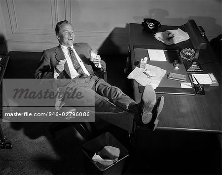 1950s SMILING MAN BUSINESSMAN SALESMAN EATING LUNCH IN OFFICE WITH FEET RESTING UP ON DESK