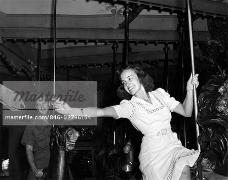 1940s WOMAN ON CAROUSEL REACHING FOR BRASS RING MAN IN BACKGROUND INDOOR