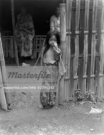 1930s LITTLE JAVANESE GIRL IN BATIK SKIRT STANDING BY BAMBOO FENCE HAND UP TO FACE JAVA INDONESIA