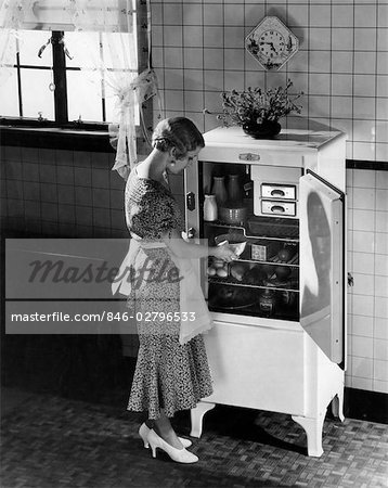 1920s 1930s BLOND WOMAN IN APRON SILK PARTY DRESS HIGH HEELS TAKING DISH FROM ELECTRIC KITCHEN REFRIGERATOR