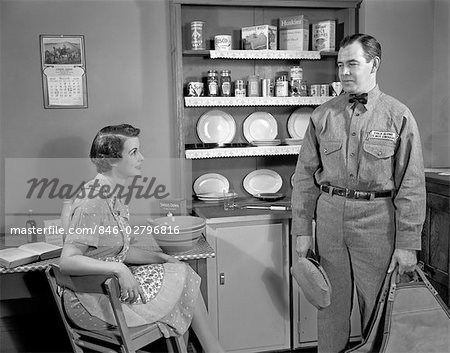 1950s WOMAN APRON SITTING AT KITCHEN TABLE TALKING TO REPAIRMAN OR HUSBAND IN UNIFORM HOUSEWIFE FOOD BOXES ON SHELVES