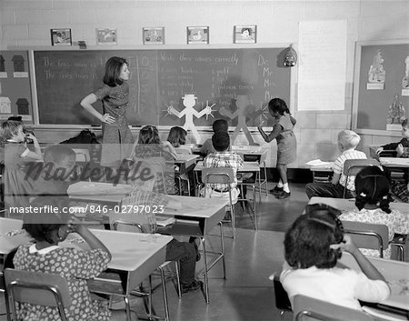 VUE DE SALLE DE CLASSE ÉLÉMENTAIRE AVEC TEACHER AT FRONT REGARDER ÉTUDIANT TRAVAILLANT SUR LE TABLEAU ARRIÈRE DES ANNÉES 1960