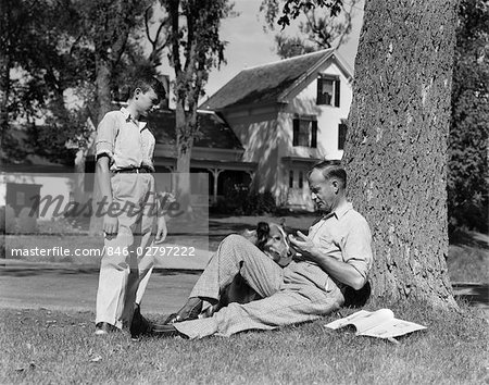 1940s FATHER AND DOG SITTING UNDER TREE TEENAGE SON STANDING FRONT YARD HOUSES STREET COMMUNITY