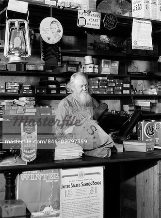1930s BEARDED GROCER BEHIND COUNTER IN GENERAL STORE LOOKING AT RUBBER BOOTS