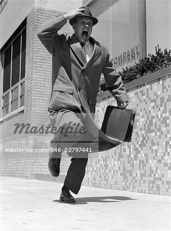 1950s FRANTIC MAN RUNNING DOWN STREET HOLDING HAT ON WITH HAND CARRYING BRIEFCASE WEARING TOP COAT COMMUTER BUSINESSMAN