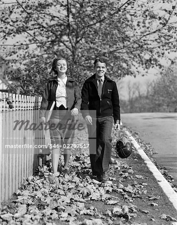1930s TEEN COUPLE WALKING ON SIDEWALK IN FALL GIRL CARRYING SCHOOLBOOKS BOY CARRYING FOOTBALL AND LEATHER HELMET SMILING OUTDOOR