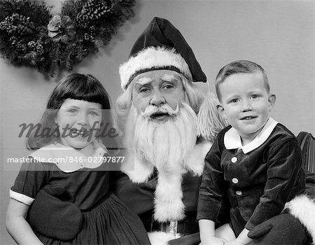 1960s MAN SANTA CLAUS POSING WITH YOUNG BOY AND GIRL ON LAP SMILING INDOOR