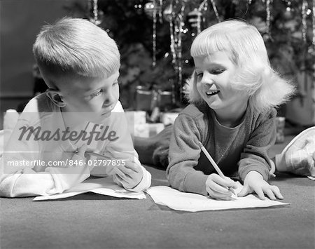 1960s BOY AND SMILING GIRL LAYING ON FLOOR EACH WITH A PENCIL AND SHEET OF PAPER WRITING CHRISTMAS TREE IN BACKGROUND