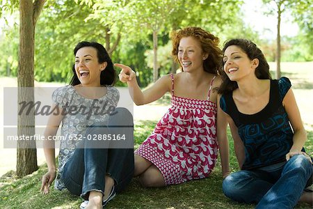 Three young women sitting in park, laughing, one pointing