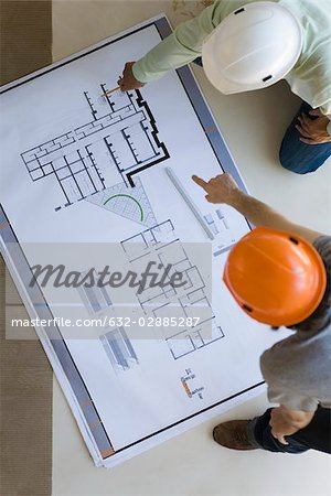 Two men crouching discussing blueprints spread out on floor