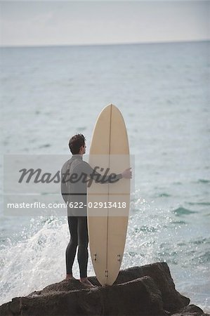 Surfer debout sur le rocher avec planche de surf à la recherche en mer