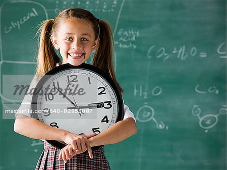 girl in a classroom standing in front of a chalkboard holding a clock