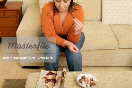 Surpoids candy jeune femme avec les cheveux bruns manger chocolat tout en regardant la photo d'une femme maigre dans un magazine