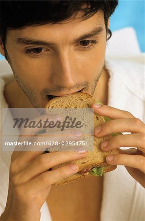 Darkhaired Man in a Bathrobe biting into a Sandwich - Snack - Foodstuff