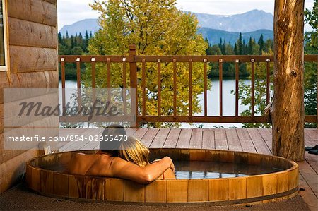 Middle aged couple relaxes in a hot tub at Winter Lake Lodge as they look out over edge of a rustic porch toward a lake.  Summer in Southcentral Alaska.