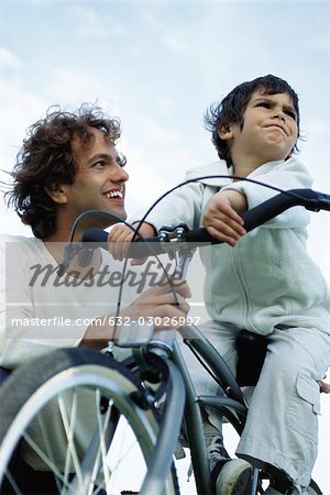 Little boy on bicycle stubbornly looking away, father kneeling at side smiling reassuringly