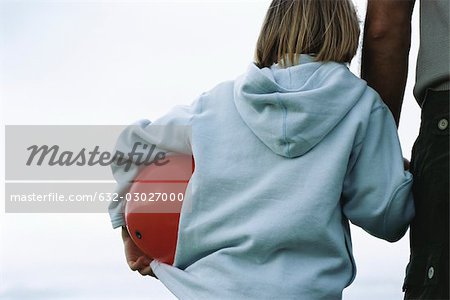 Young boy holding father's hand, carrying ball under arm, rear view