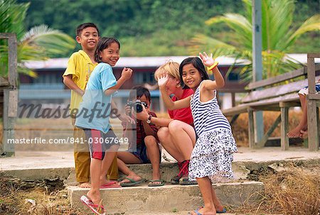 A group of Iban Dayak children with a tourist and camera outside their longhouse on the Rajang River near Kapit in Sarawak, northwest Borneo, Malaysia, Southeast Asia, Asia