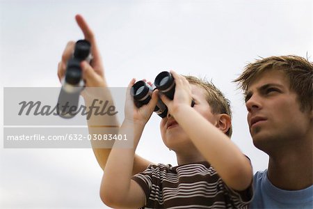 Father and son using binoculars, man pointing skyward