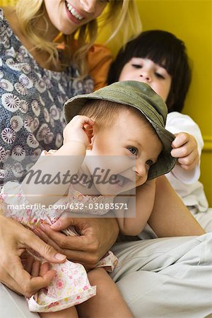 Infant girl sitting with mother and brother, wearing hat and laughing