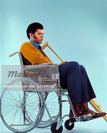 1970s SAD AFRICAN AMERICAN MAN SITTING IN WHEELCHAIR HOLDING CRUTCHES