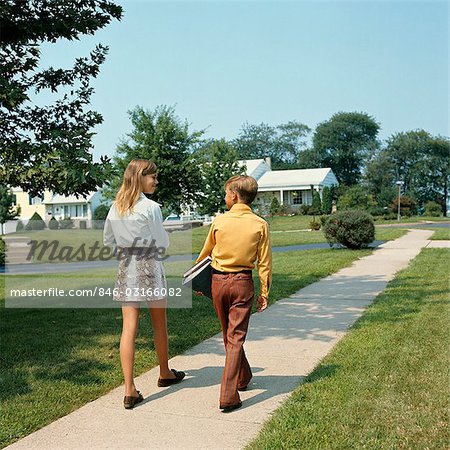 ANNÉES 1970 ADO GARÇON ET FILLE MARCHER DANS LA RUE DE BANLIEUE TRANSPORTANT LES MANUELS SCOLAIRES