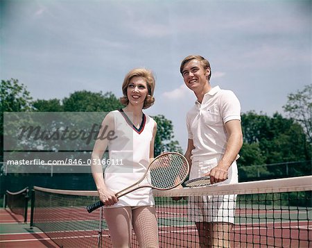 1970s SMILING COUPLE STANDING ON OPPOSITE SIDES OF TENNIS COURT NET HOLDING RACKETS