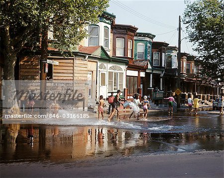 1970s CHILDREN IN URBAN NEIGHBORHOOD PLAYING IN STREET IN WATER SPRAY FROM HYDRANT