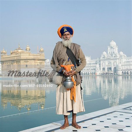 Sikh man in traditional clothing standing near a pond with a temple in the background, Golden Temple, Amritsar, Punjab, India