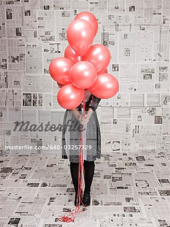 Young woman hiding face behind bunch of balloons in newspapers covered room, studio shot
