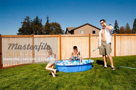 Man with hose talking on cell phone spraying boy with hose in children's pool