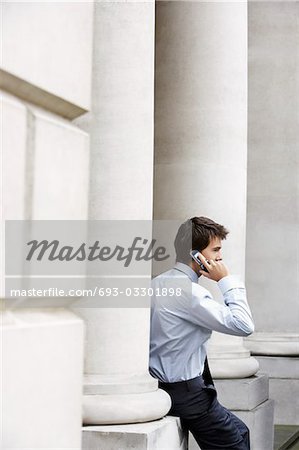 Businessman on cell phone outside building sitting on pillar