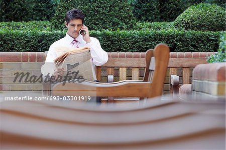 Businessman on park bench reading newspaper and talking on cell phone