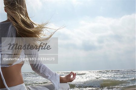 Jeune femme méditant sur la plage, face à l'océan, vue arrière