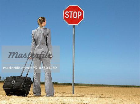 Business woman with luggage, standing in front of stop sign in desert, back view