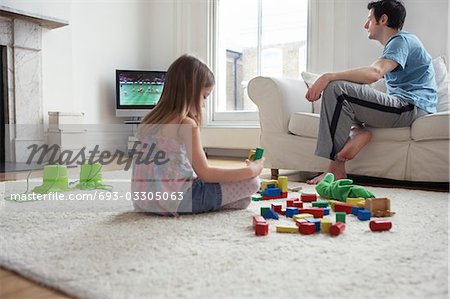 Girl (5-6) sitting on floor, playing with blocks, father watching television