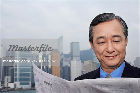 Middle-aged business man reading newspaper, office buildings in background