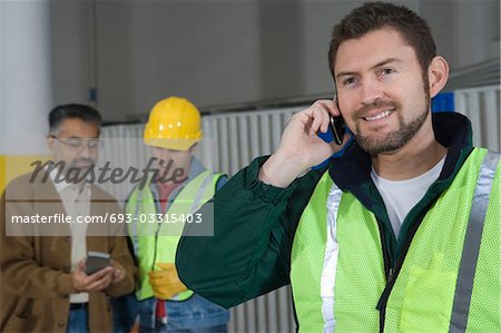 Homme debout en usine, à l'aide de téléphone, ses collègues en arrière-plan