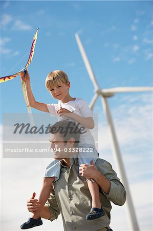 Boy (7-9) holding kite, sitting on fathers shoulders at wind farm