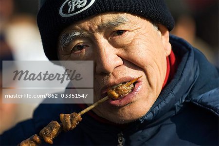 Chine, Beijing. Chinese New année Spring Festival - Changdian rue juste - un vieil homme manger shish kebab.