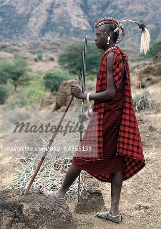 A Maasai warrior with his hair styled in a most unusual way. His long braids have been wrapped tightly in leather,decorated with beads and tied in an arch over his head. A colobus monkey tail sets this singular hairstyle apart from the more traditional warrior styles.